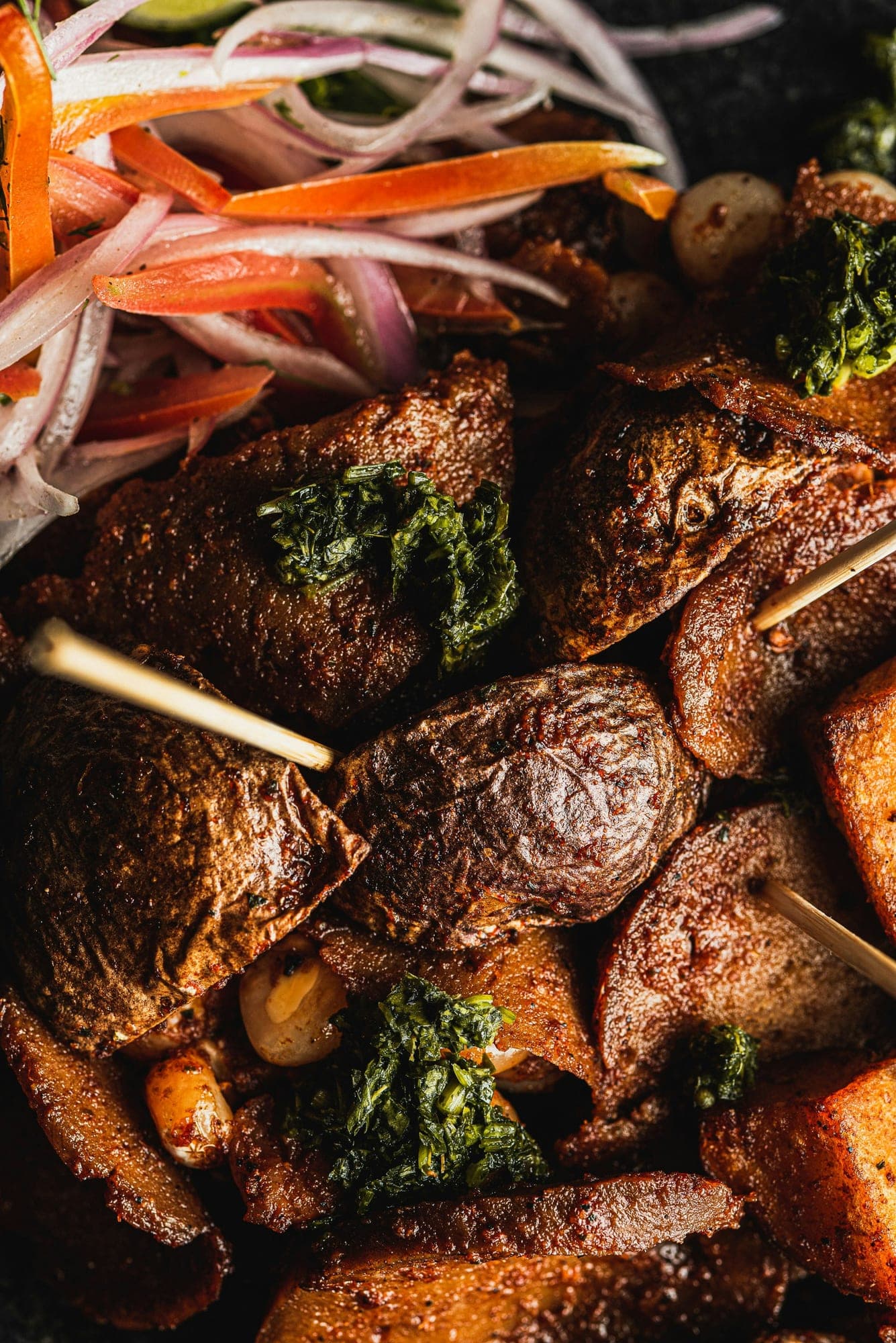 Overhead close-up of grilled suya on wooden skewers, crusted in yaji and topped with chopped herbs, with sliced red onion, tomato and cucumber piled alongside.